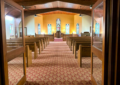 The entrance of the chapel with the alter in the back. The chapel room features a beautiful curved wooden ceiling supported by large beams. The pews are in a beautiful oak wood brown.