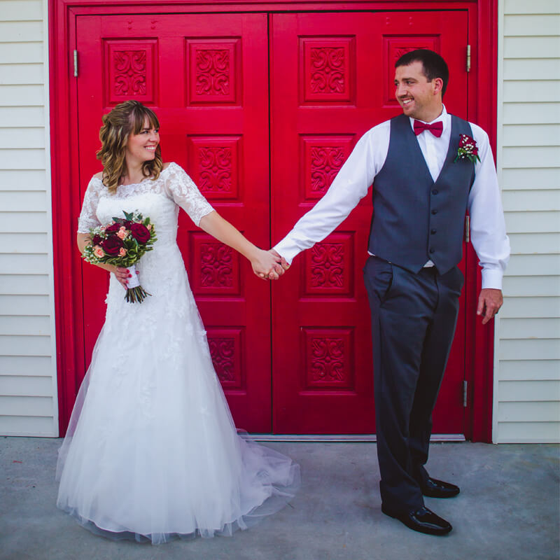 SYCAMORE-SPRINGS-PARK-WEDDING-1 SYCAMORE-SPRINGS-PARK-WEDDING-A couple is standing in from of the beautiful red doors that lead into the chapel. The bride is in her white wedding gown holding a bouquet of flowers in one hand and hold her grooms hand in the other. He is wearing a suit and both are looking back over their shoulder at each other.